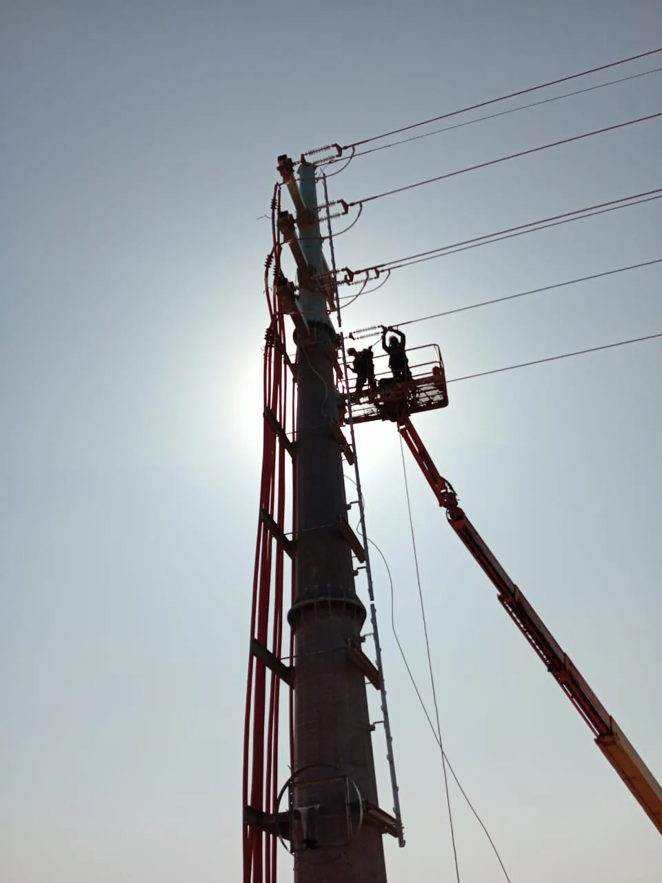 Deux techniciens en nacelle élévatrice raccordent les isolateurs en tête d’un pylône haute tension, à contre-jour en fin de journée
